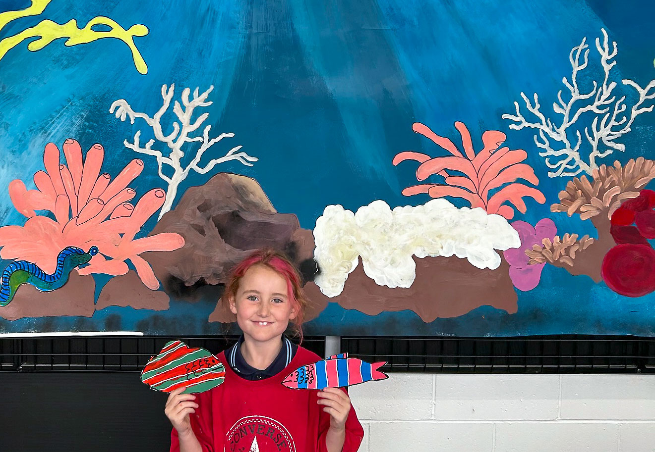 Under the Sea - collaborative installation by the Upstart kids. Young artist holding a brightly coloured fish against an painted undersea backdrop.
