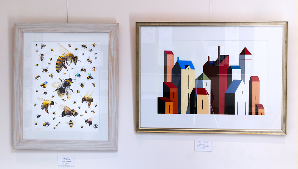 Framed artworks by Joan Denner, one of bees in watercolour and one of stylized buildings in bright colours.
