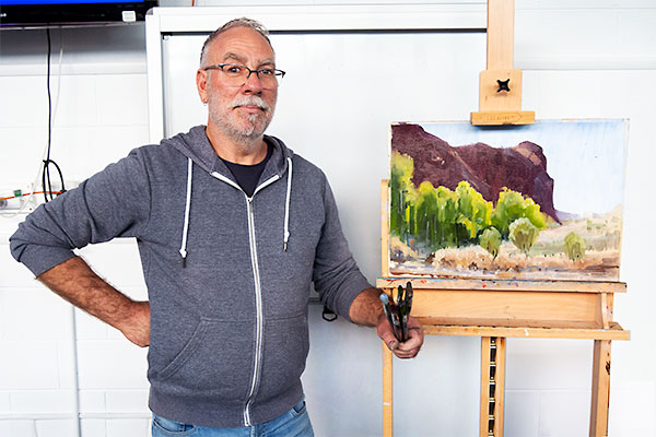 Ben Winspear with completed demo painting of a landscape with rocky hill and trees in foreground.