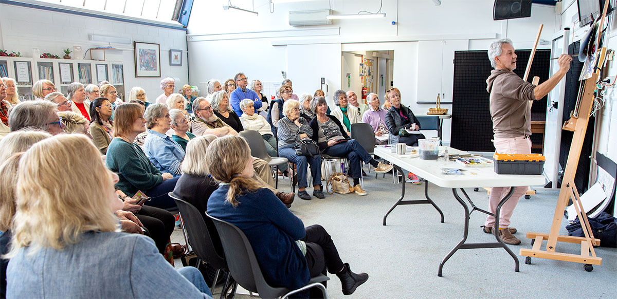 Greg Allen Demo at Peninsula Arts Society. Greg painting at an easel with a large group of seated people watching.